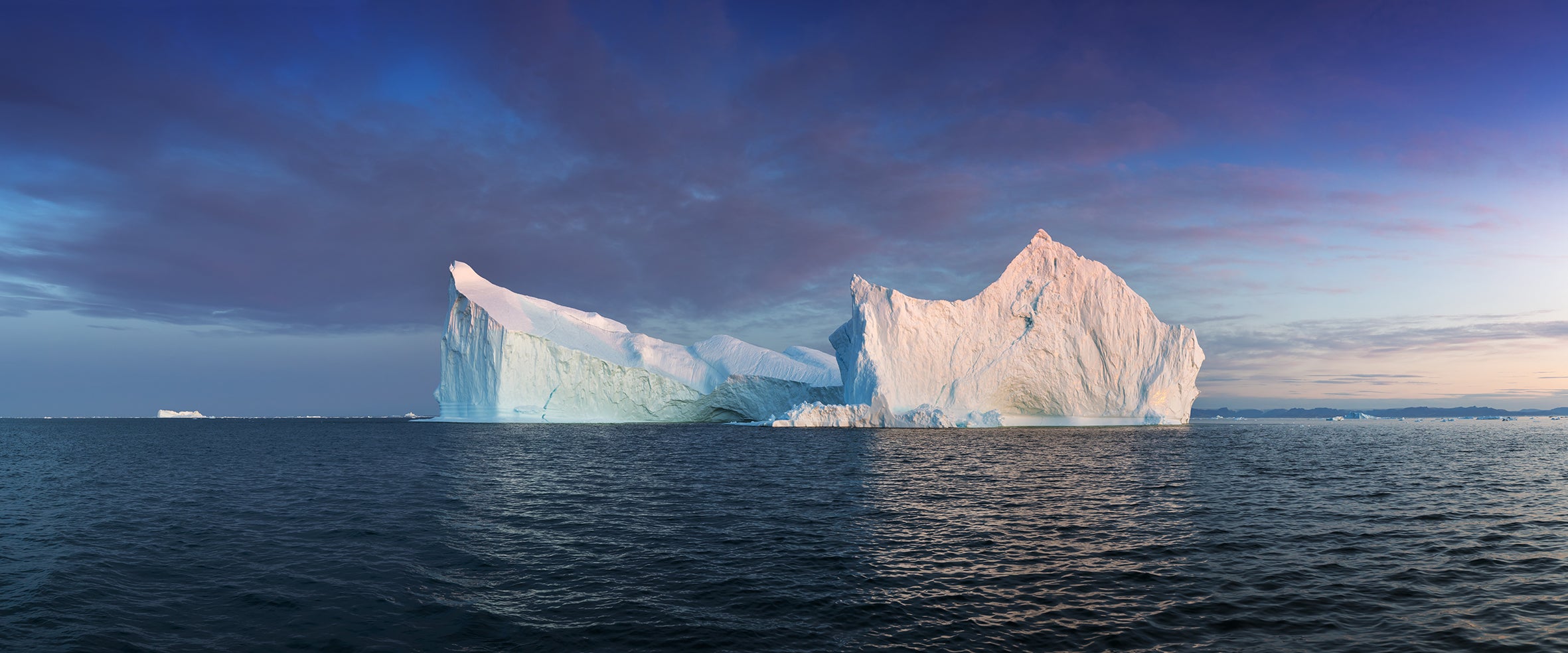 Two large icebergs floating in the ocean with a blue sky and clouds in the background.