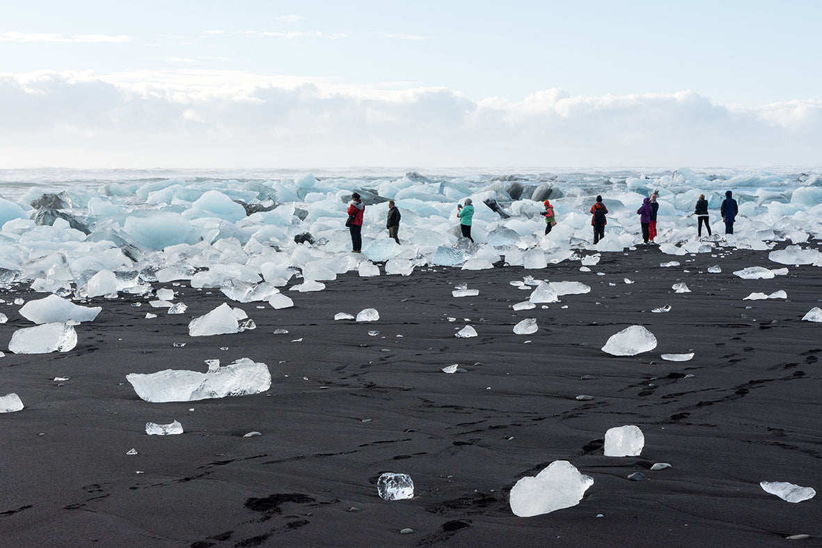 People standing on a black sand beach with large ice blocks and a glacier in the background.