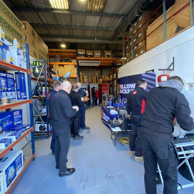 Group of people in a warehouse setting with equipment and storage shelves.