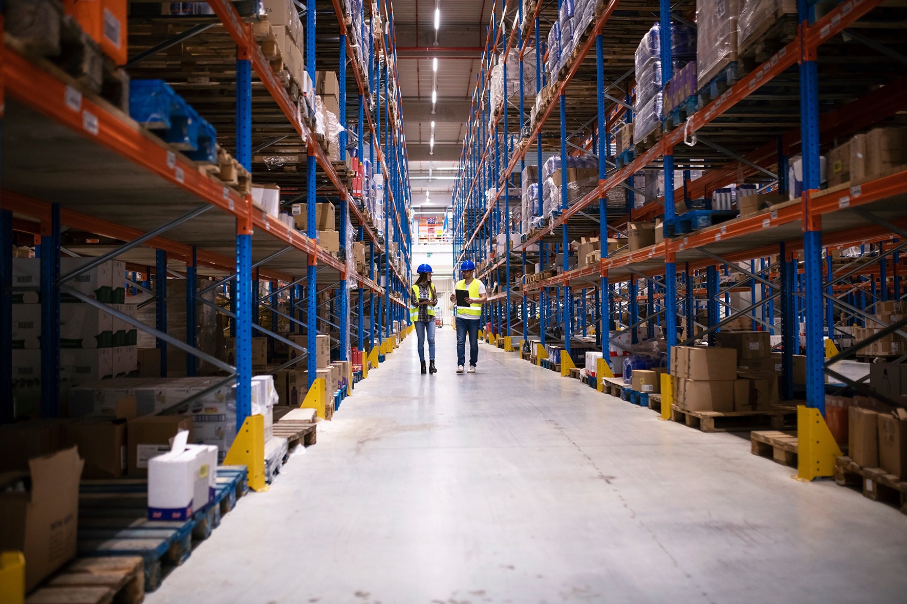 Two workers in a warehouse with high shelves and boxes.