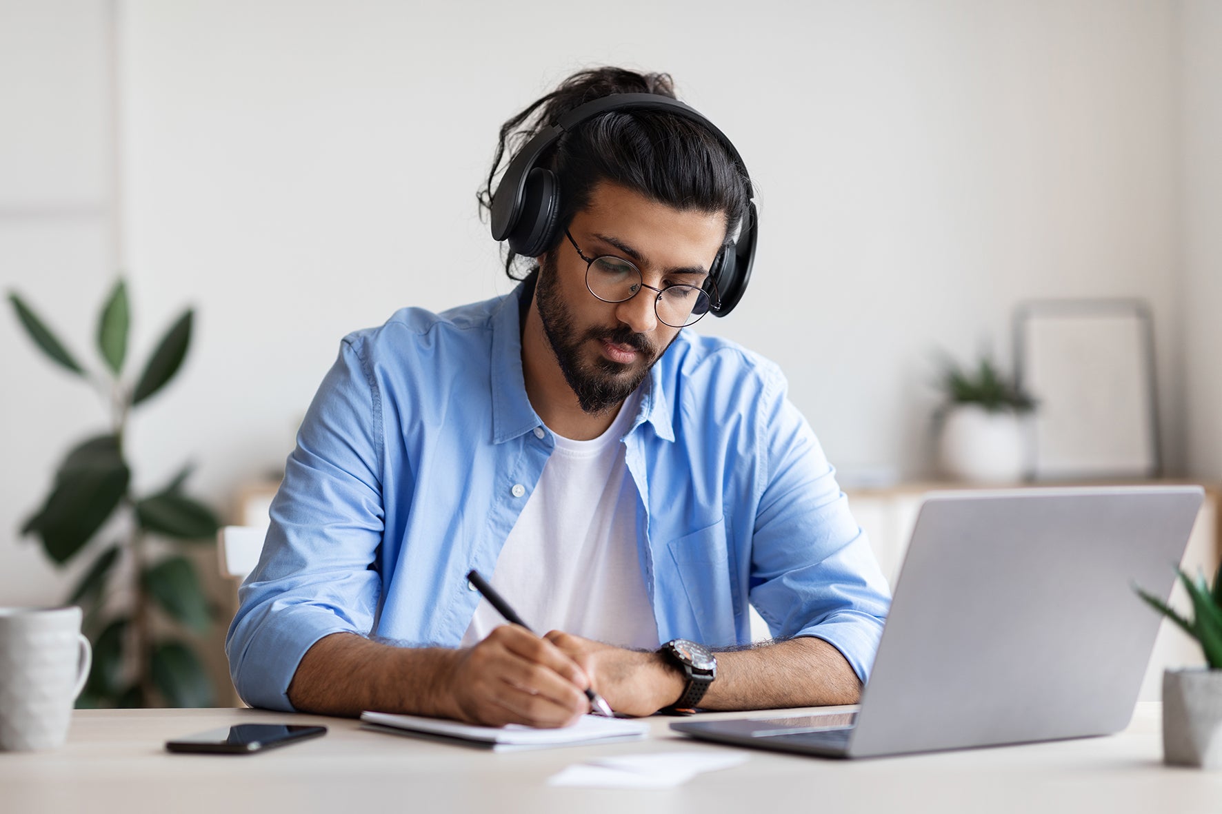 Man wearing headphones and working on a laptop with a notebook and pen at a desk.