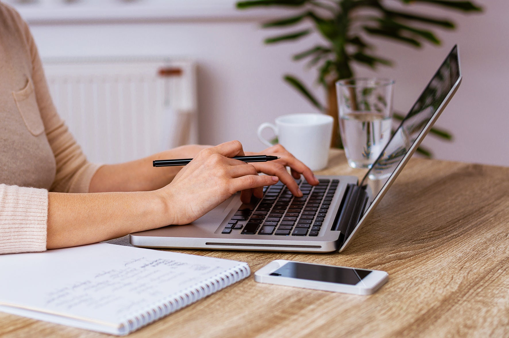 Person using a laptop on a wooden desk with a notebook, smartphone, and coffee cup.