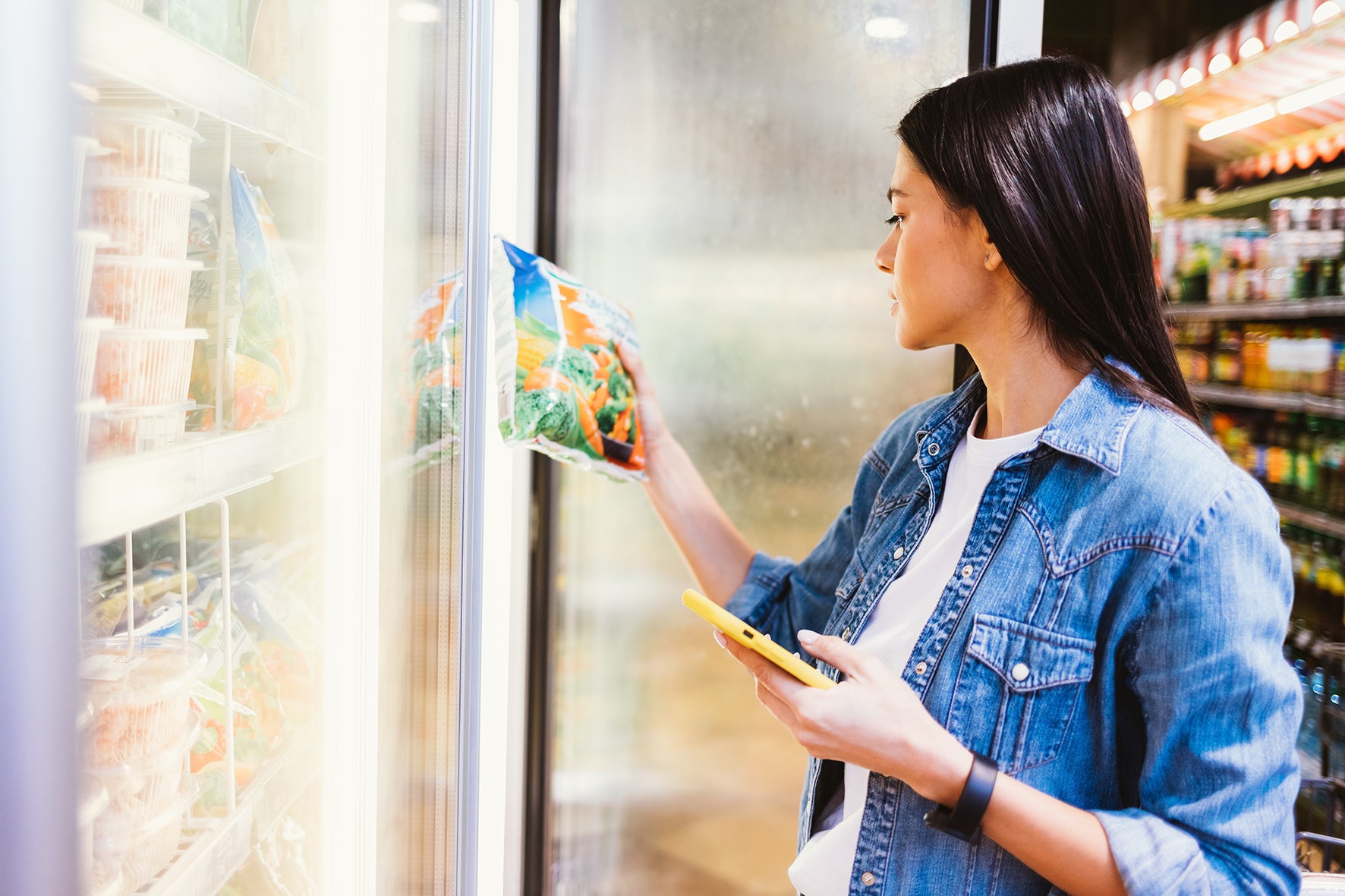 A woman in a blue denim jacket stands in a grocery store freezer aisle, holding a bag of frozen vegetables next to a glass door while looking at her phone.