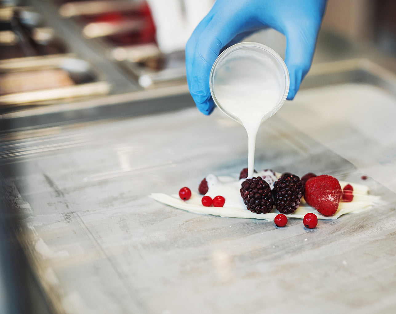 Hand in blue glove pouring white cream over berries on a metal surface