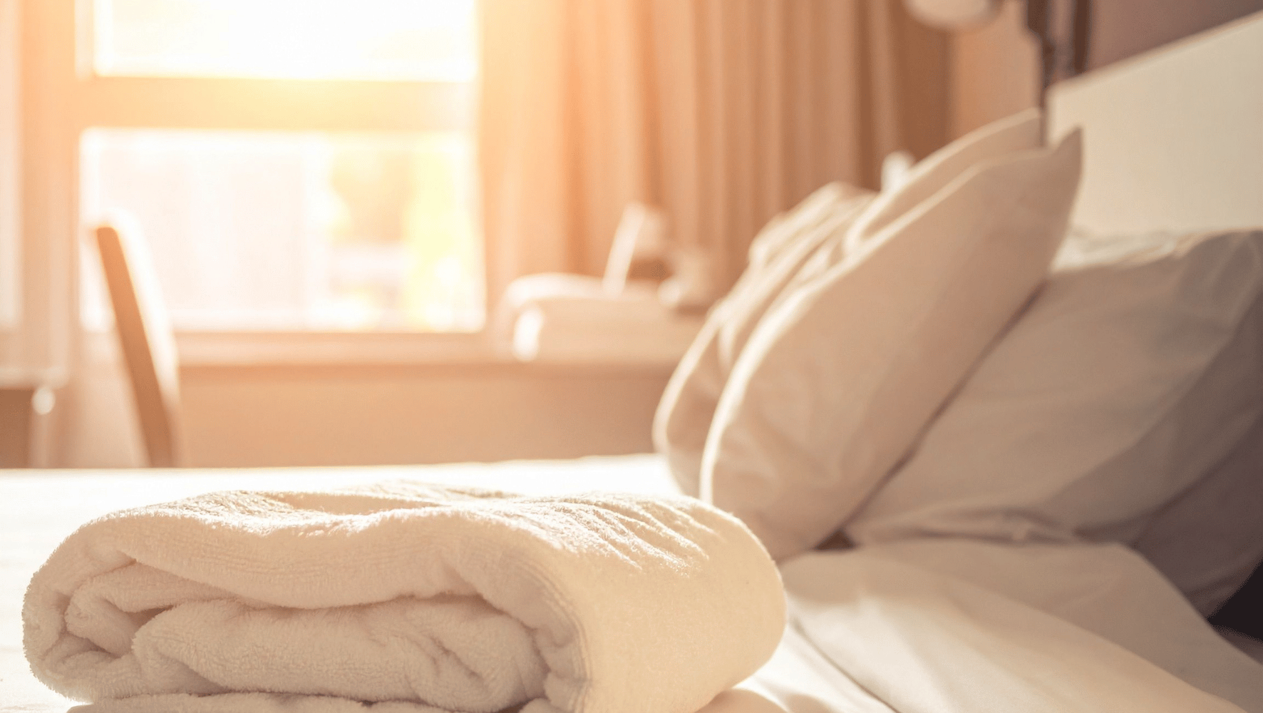 A neatly folded cream-colored blanket or towel rests on a bed with matching white sheets and pillows in a softly lit bedroom. Warm sunlight streams in from a window in the background.