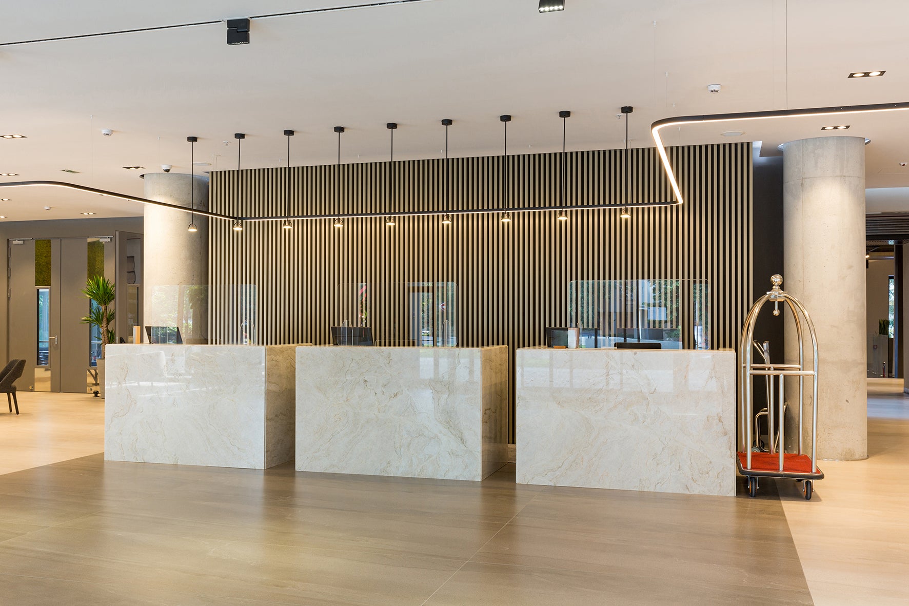 A modern hotel lobby reception area with three marble reception desks, a slatted wood back wall with warm lighting, a gold luggage cart, and light stone flooring.