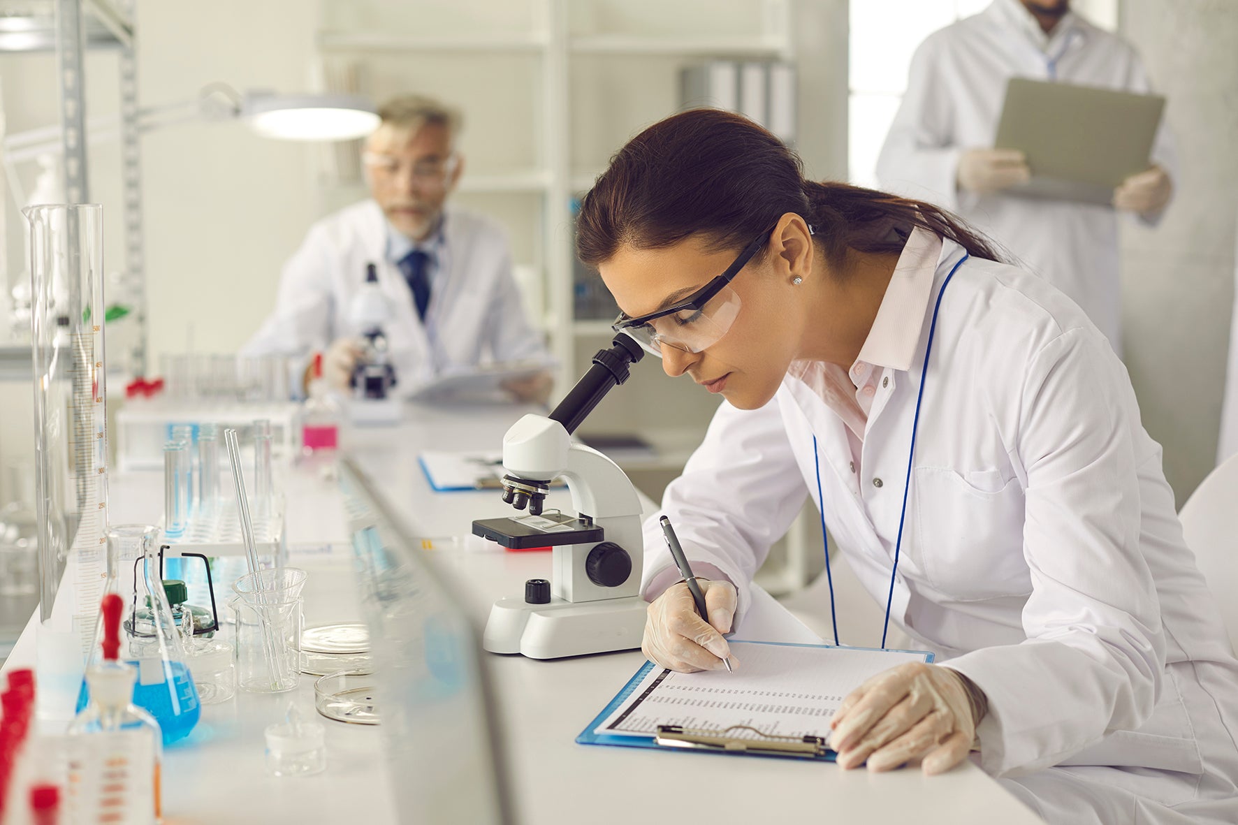 A woman in a white lab coat looking through microscope in a laboratory, with a clipboard.