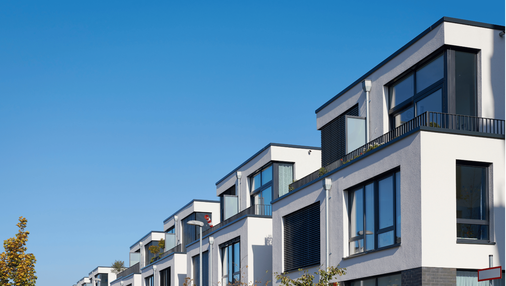 Modern multi-story residential buildings with white and gray facades, dark window frames, and balconies under a clear blue sky.