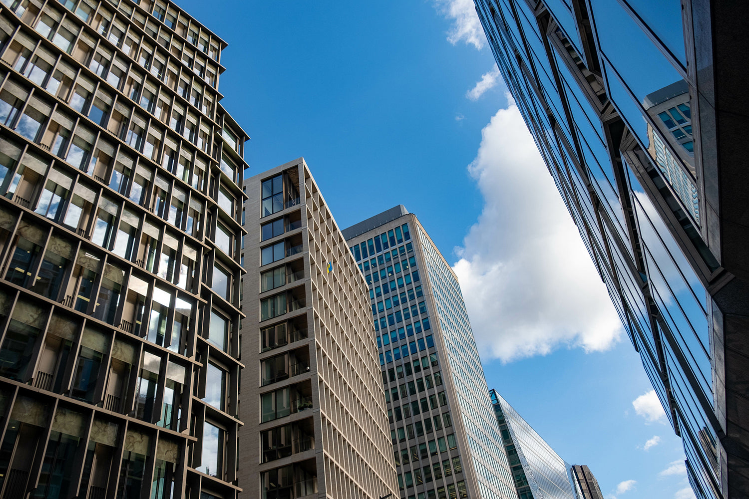 Low-angle view of modern skyscrapers with glass and concrete facades against a bright blue sky with white clouds.