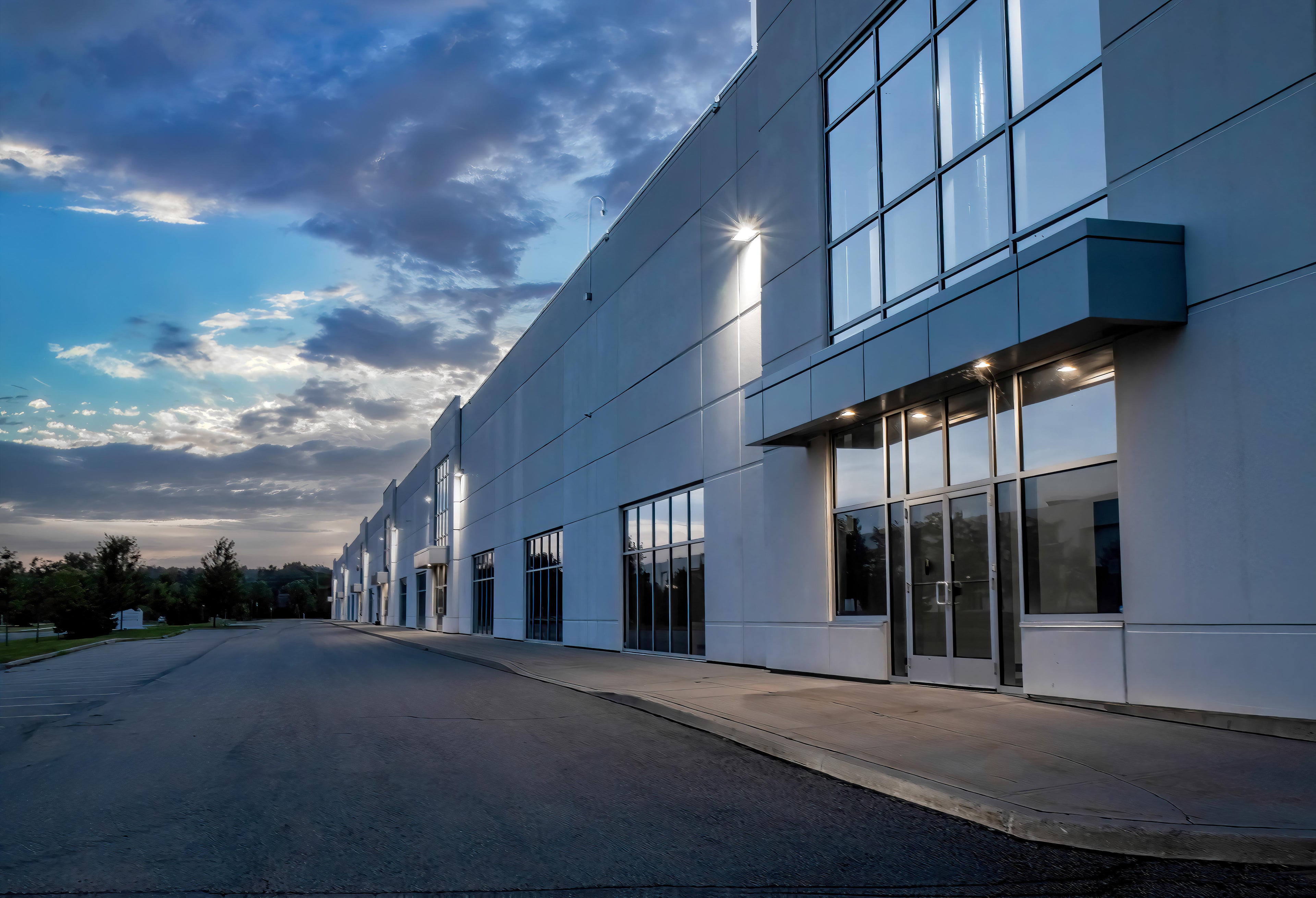 A long, modern white commercial building with large glass windows and a paved driveway, photographed at dusk under a cloudy blue sky.