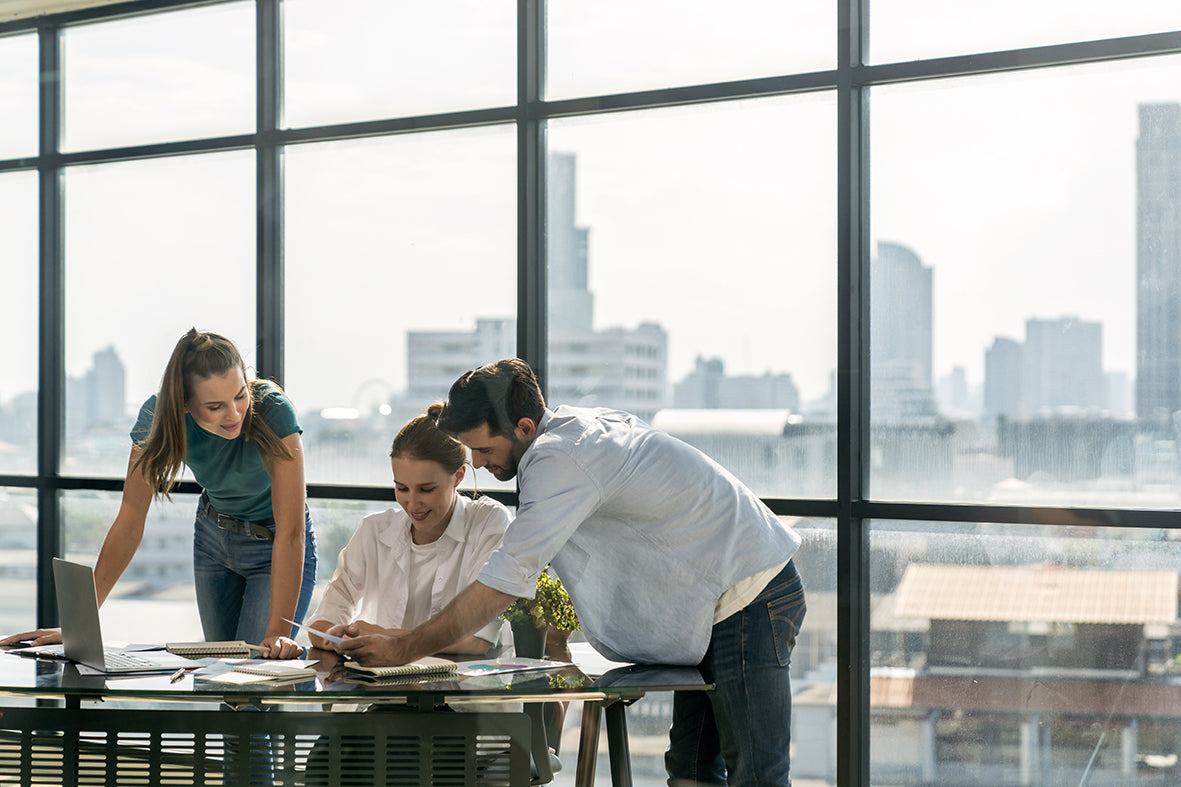 Three colleagues in a modern office with large floor-to-ceiling windows, collaborating around a desk with a laptop and documents. The background shows a blurred city skyline.