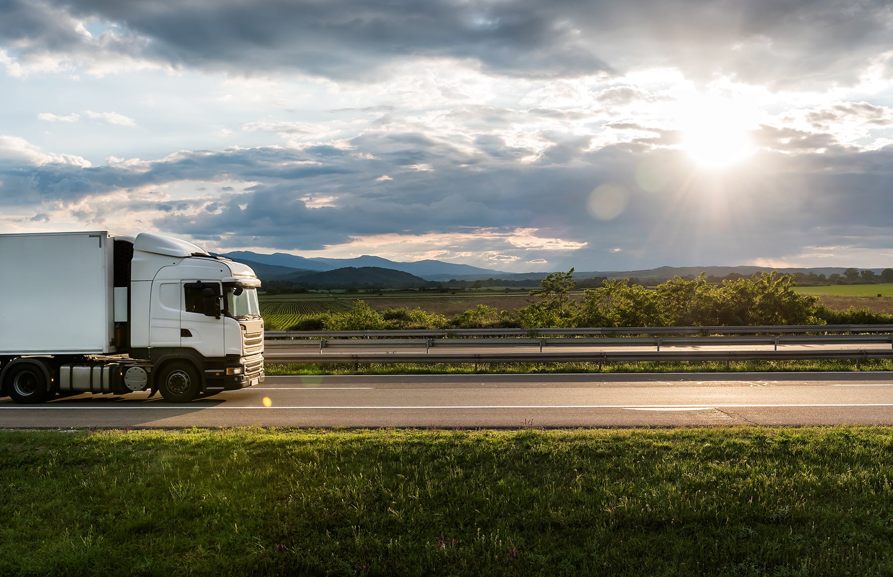A white box truck drives along a highway beside grassy fields and distant mountains under a dramatic cloudy sky with bright sun rays.