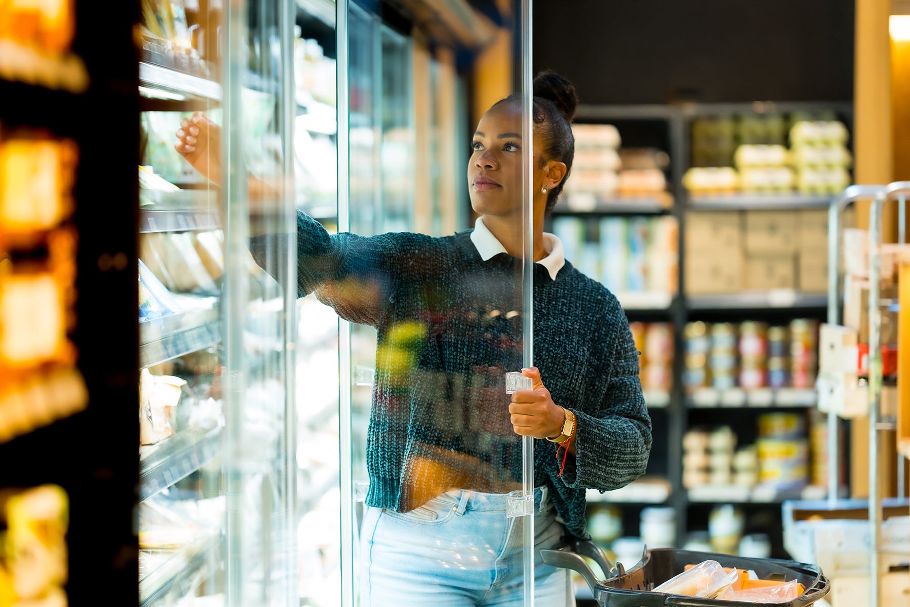 A woman in a dark knit sweater and light blue jeans reaches into a glass-front refrigerator inside a grocery store aisle, with shelves of packaged goods blurred in the background.